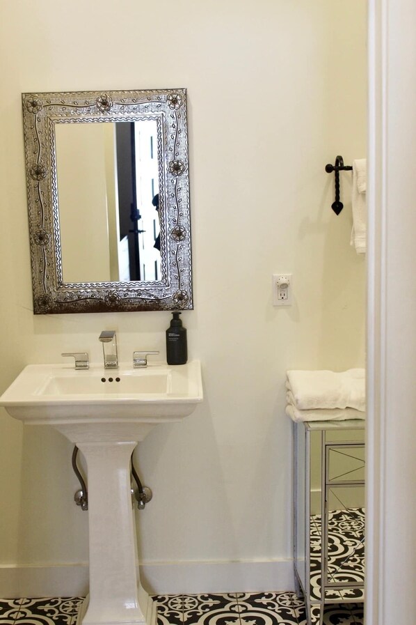 A bathroom with a white pedestal sink, a mirror with a decorative frame, a black bottle of shampoo, and patterned floor tiles. - Lodge 1 on the Mason square (Mason)