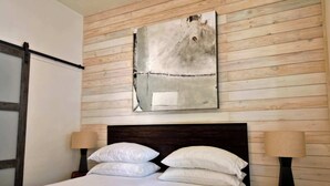 Une chambre avec un mur recouvert de lambris en bois, une grande peinture abstraite, une tête de lit sombre, deux lampes de chevet et un lit bien fait, avec du linge de lit blanc.