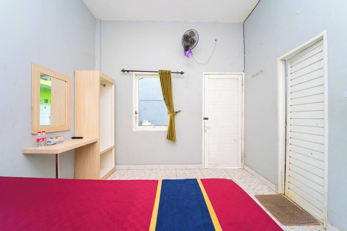 A bedroom with a bed, a desk, a wardrobe, a window with curtains, a door, and a fan.