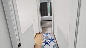 A narrow hallway with white walls, a tiled floor, and a patterned rug leading to a room with a bed and a window.