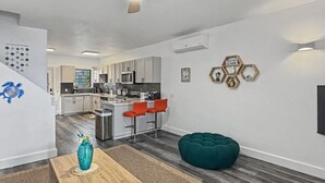 A modern kitchen with white cabinets, a microwave, and a coffee machine. There is a dining area with a wooden table and orange chairs. A green ottoman and a blue vase are on the floor.