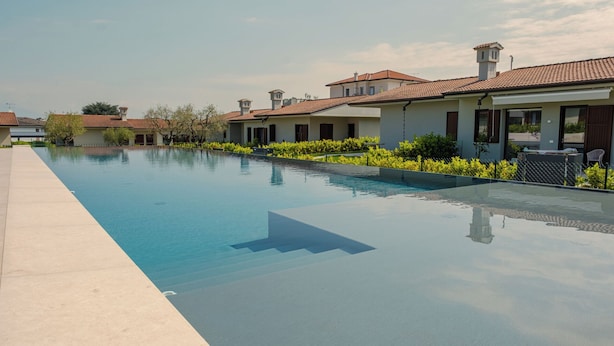 Una piscina con superficie azul cristalina, rodeada por un seto bajo y una hilera de casas con tejados de tejas.