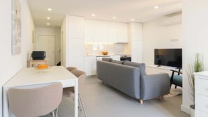 Een moderne woonkamer met een grijze bank, een eettafel met stoelen, een televisie en een keuken met witte kasten en een marmeren achterwand.