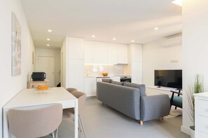 Un soggiorno moderno con un divano grigio, un tavolo da pranzo con sedie, una TV e un angolo cottura con pensili bianchi e una parete backsplash in marmo.