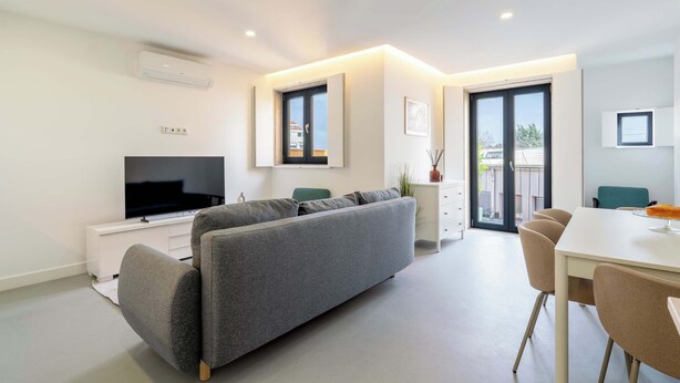 Een moderne woonkamer met een grijze bank, een flatscreen televisie, een eethoek met een tafel en stoelen, en een balkon met uitzicht.