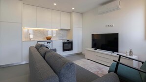 Een moderne keuken met witte kasten, een marmeren aanrechtblad en een ingebouwde oven. In de woonkamer staat een grijze bank en een groene fauteuil, en er is een flatscreen televisie aan de wand bevestigd.