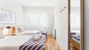 Une chambre d’hôtel avec deux lits, une grande fenêtre avec des rideaux légers, un radiateur et un miroir au mur.