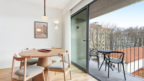 Un espace repas moderne avec une table ronde en bois et des chaises, un luminaire suspendu et une grande porte vitrée donnant sur un balcon avec vue sur les bâtiments et les arbres.
