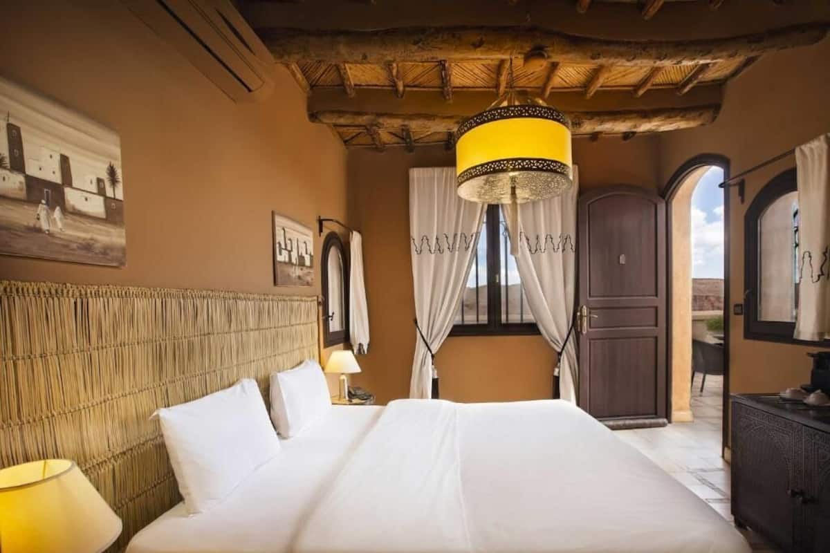 A hotel room with a large bed, a wooden headboard, a chandelier, and a view through an open door.