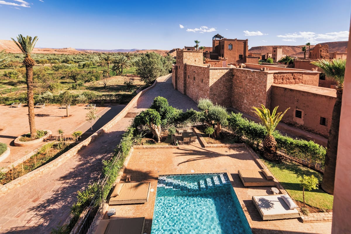 A pool area with lounge chairs, a clear blue pool, and a view of traditional buildings and greenery.