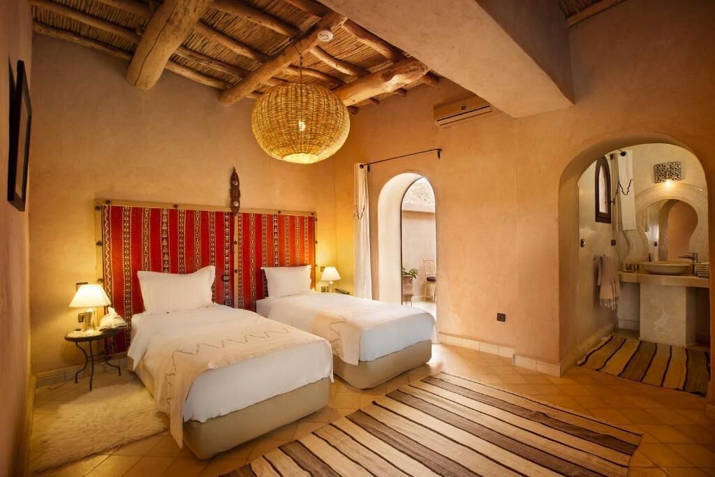 A room with two beds, a woven ceiling light, a red curtain, and a small bathroom with a sink and mirror.