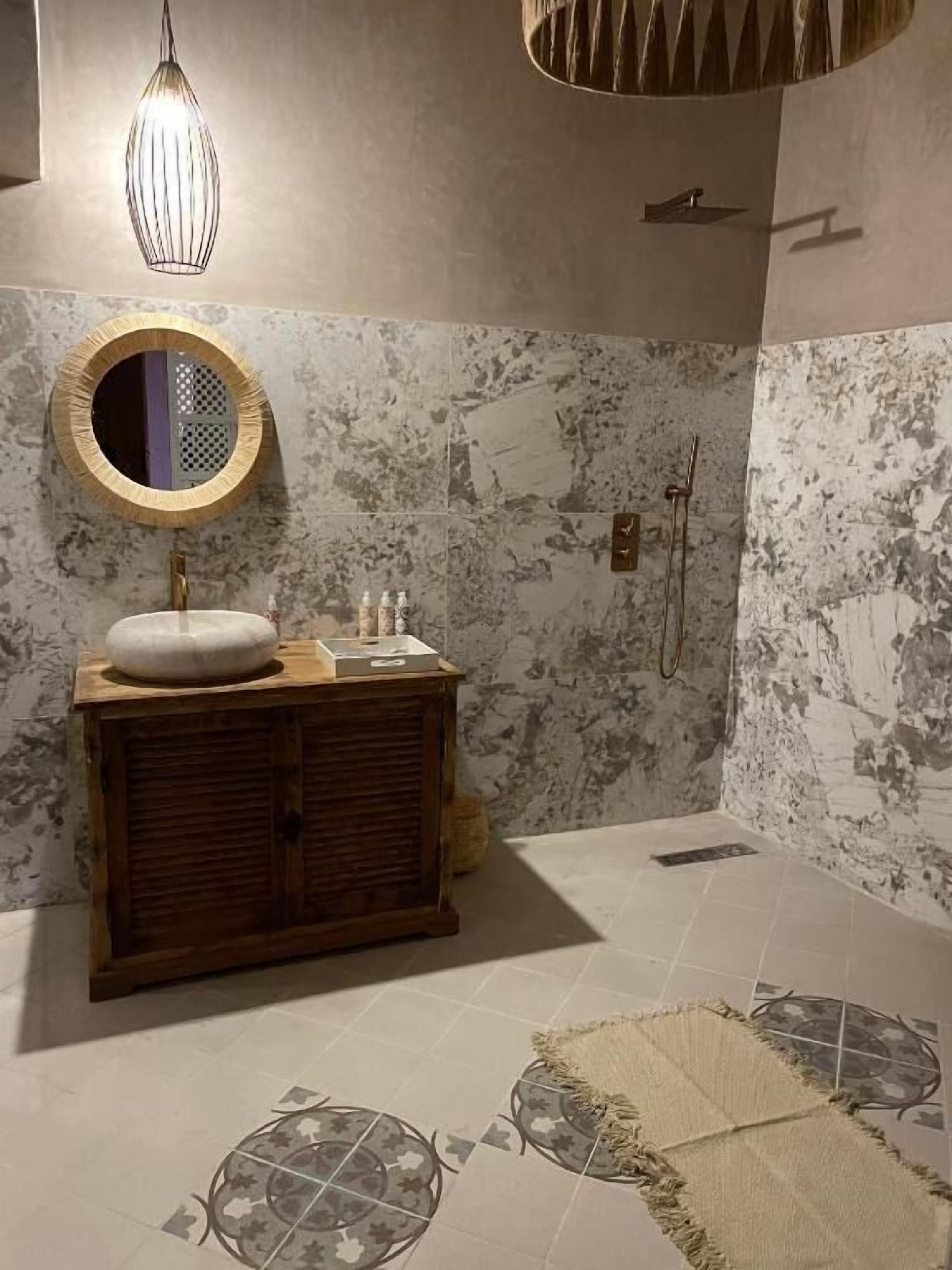 A bathroom with a marble wall, a wooden vanity, a round mirror, a hanging light fixture, and a shower area.