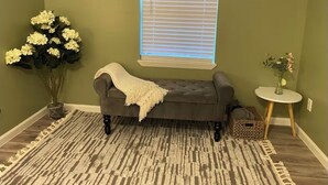 Ein Wohnzimmer mit grauem Sofa, einem gemusterten Teppich, einer Vase mit Blumen und einem Fenster mit Jalousien.
