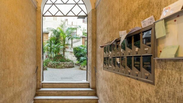 A corridor with a metal mail slot on the right wall and a view of a courtyard with plants and buildings outside. - ZUCCHELLI ROMAN RESIDENCE (Roma)