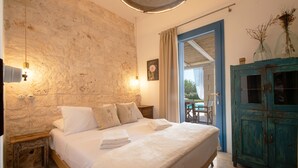 Ein Schlafzimmer mit einer Steinwand, einem Bett mit weißer Bettwäsche, einem hölzernen Nachttisch, einem blauen Schrank und Blick ins Freie.