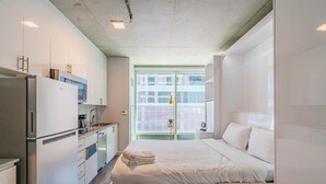 Une chambre d’hôtel moderne dotée d’une kitchenette, d’un lit avec du linge de maison blanc et offrant une vue sur la ville grâce à de grandes fenêtres.