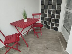 Een rode metalen tafel en stoelen staan op een balkon met uitzicht op een rijtje ramen en een bloempot.