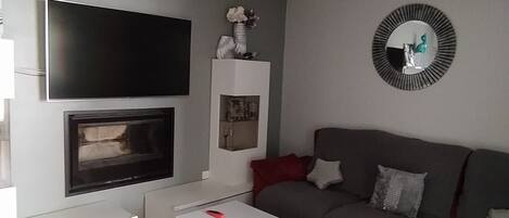 A modern living room with a flat-screen TV, fireplace, white coffee table, and a grey sofa with red cushions.