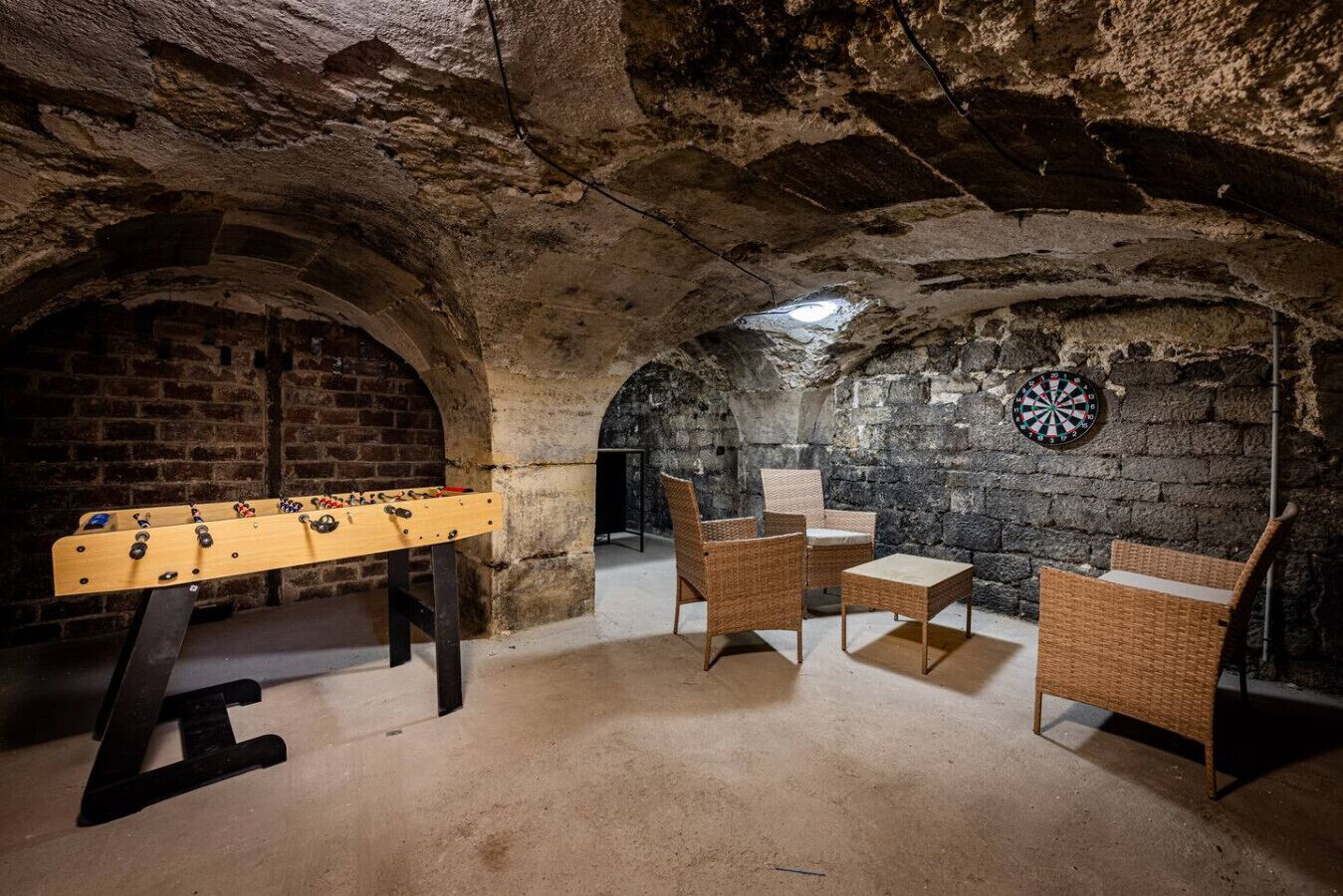 A room with a foosball table, wicker chairs, and a dartboard.