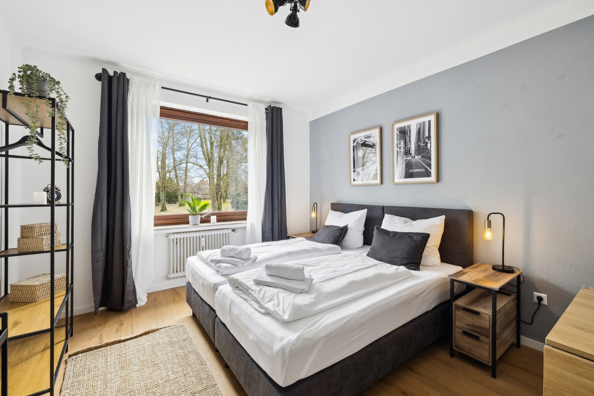 Un dormitorio moderno con una cama grande, una mesita de noche, un radiador, una ventana con cortinas, una planta en un estante y dos cuadros en la pared.