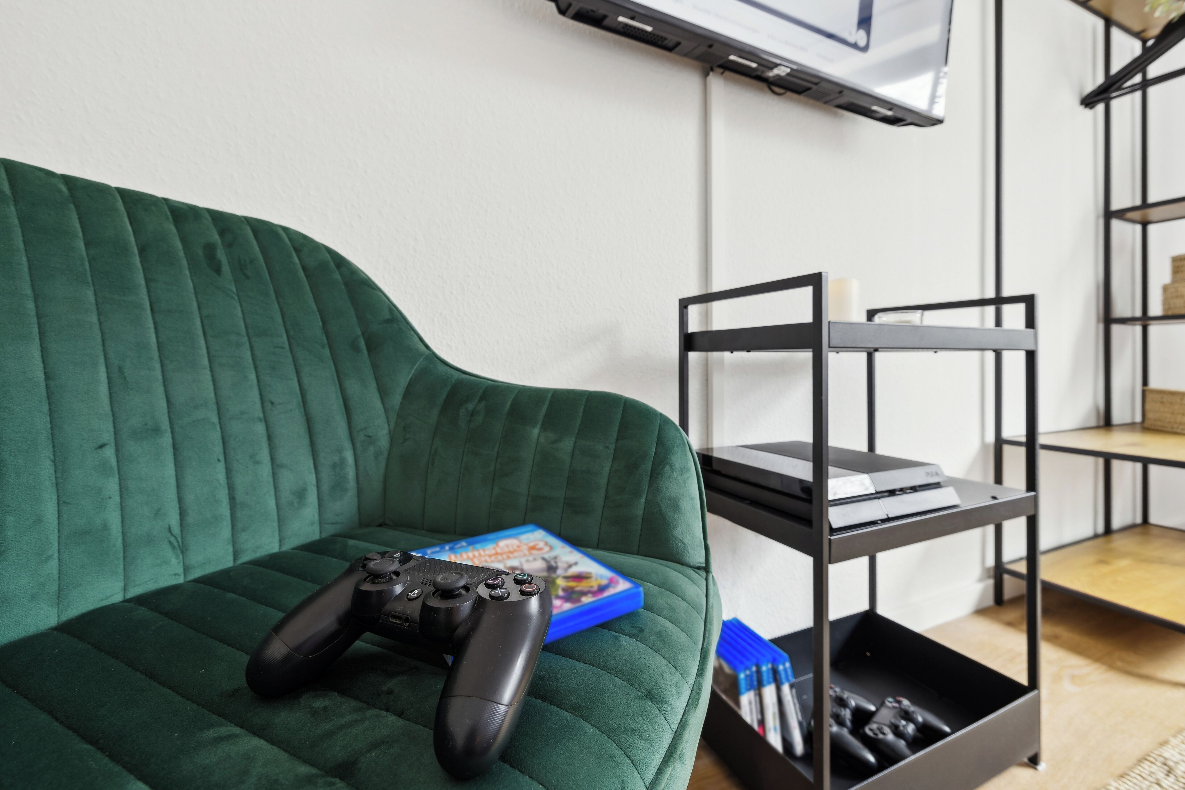 Ein grünes Sofa mit Spielregler und Spielehülle, ein Metallregal und ein an der Wand befestigter Fernseher.