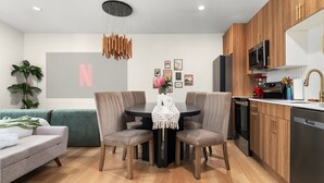 A modern kitchen with wooden cabinets, a built-in oven, and a sink. There is a dining area with a round table and chairs, and a sofa in the living room. - Zen Den · Hot Tub · Cold Plunge · Movie Area (Austin)