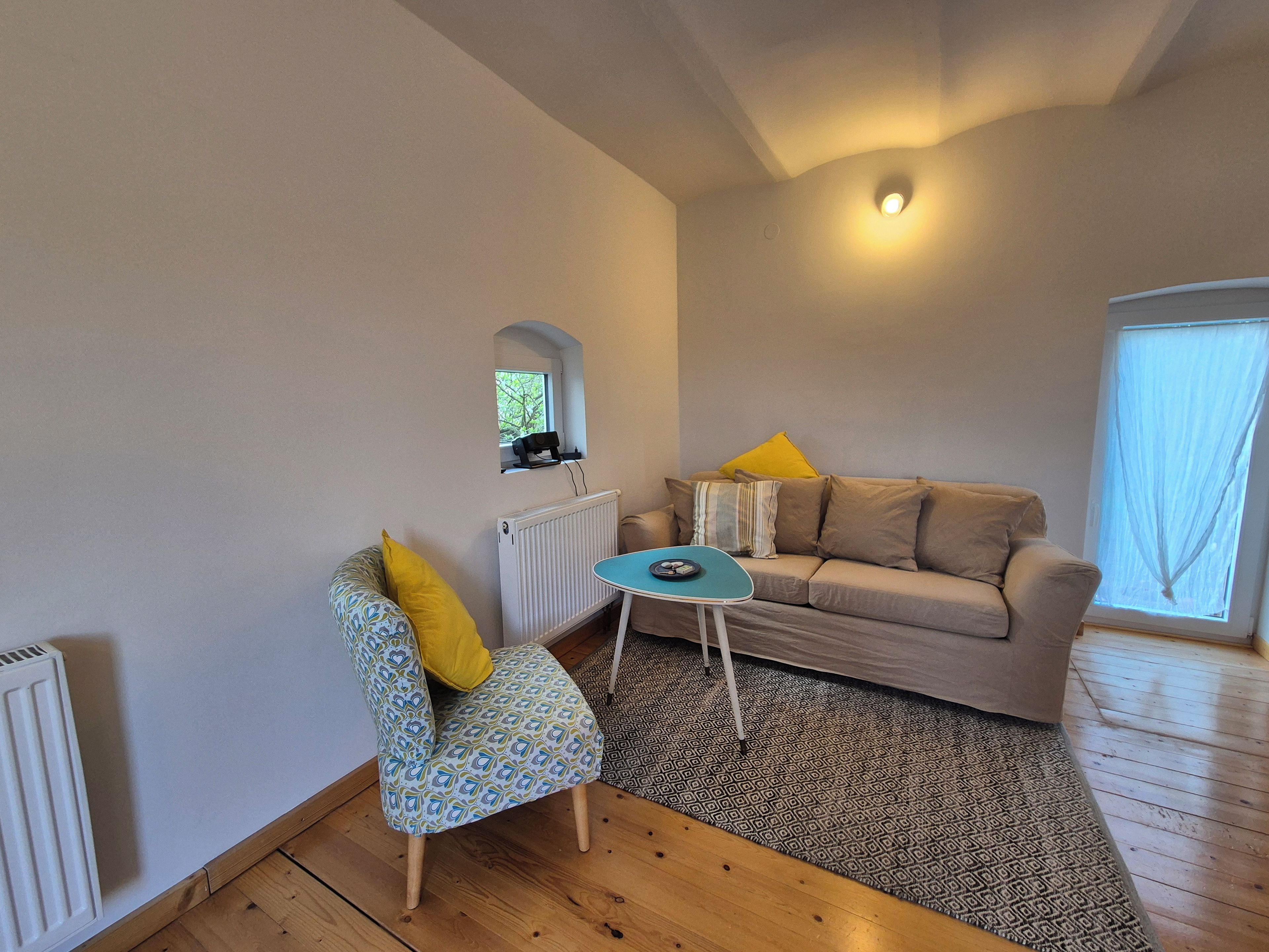 Ein Wohnzimmer mit einer Couch, einem gemusterten Sessel, einem kleinen Tisch, einer Heizung und einem Fenster mit Vorhängen.