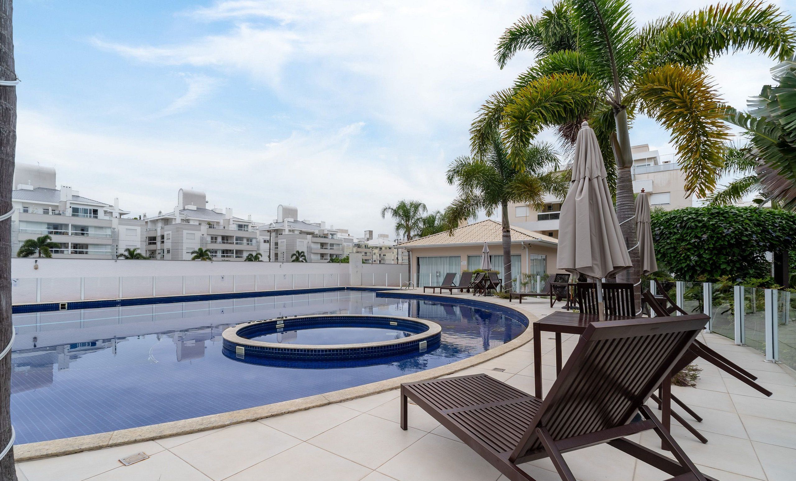 A rooftop swimming pool with lounge chairs and umbrellas, surrounded by palm trees and modern buildings.