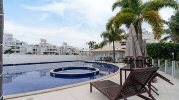 A rooftop swimming pool with lounge chairs and umbrellas, surrounded by palm trees and modern buildings.