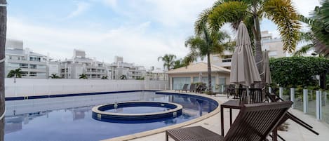 Una piscina en la azotea con sillas de descanso y sombrillas, rodeada de palmeras y edificios modernos.