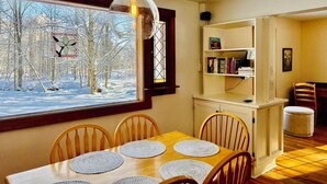 Een eetruimte met een houten tafel en stoelen, een raam met uitzicht op een besneeuwd bos, en een boekenkast op de achtergrond.