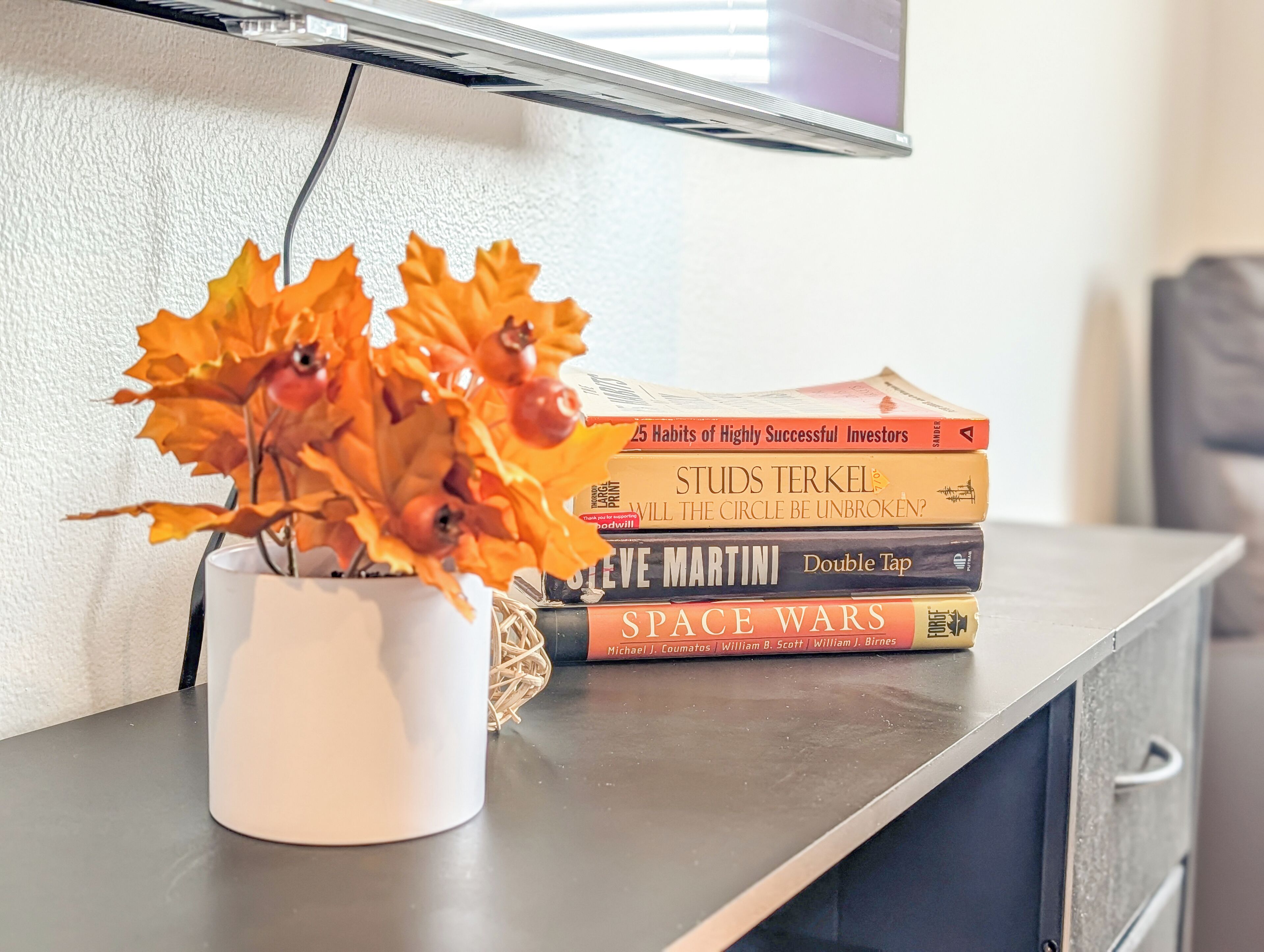 A white vase with orange leaves and berries on a table, next to a stack of books including '25 Habits of Highly Successful Investors' and 'Space Wars.'