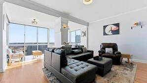 A modern living room with a large sectional sofa, a leather armchair, and a small table. There is a large window offering a view of the city. - The ATL Sky Suite High-Floor 3BR Downtown Penthouse Stunning City Views (Atlanta)