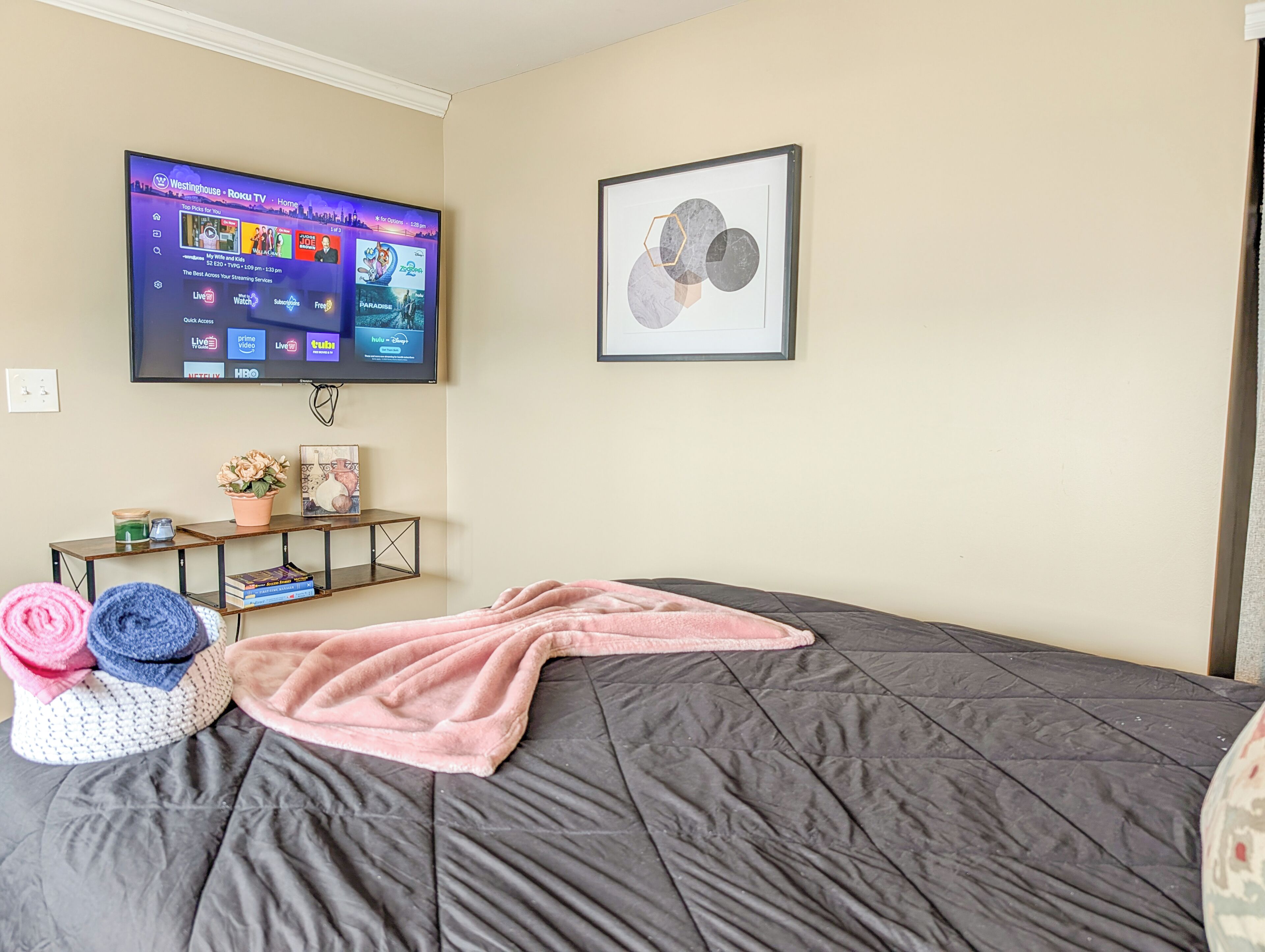 Ein Schlafzimmer mit einem Bett, einem an der Wand montierten Fernseher, einem gerahmten Bild und einem kleinen Tisch mit Pflanze und Büchern.
