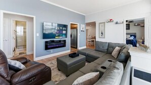 A modern living room with a large flat-screen TV mounted on the wall, a grey sectional sofa, a leather armchair, and a small ottoman. - The ATL Sky Suite High-Floor 3BR Downtown Penthouse Stunning City Views (Atlanta)