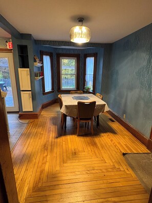 A dining room with a wooden herringbone floor, a round table with chairs, a chandelier, and windows with curtains. - Charming 4-bedroom Victorian home in Milton!  Close to I-89, the lake, & skiing! (Milton)