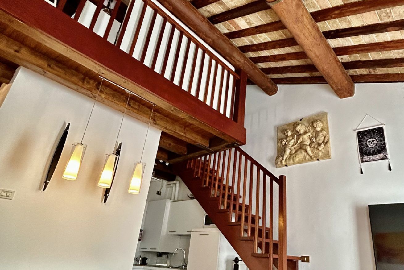 Un escalier avec des rampes en bois menant à un niveau supérieur, un coin cuisine à l’arrière-plan et une œuvre d’art accrochée au mur.