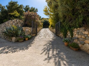 A gravel pathway leading to a gated entrance, flanked by stone walls and various potted plants. - Villa Claudia Relax & Garden – Torre delle Stelle (Torre Delle Stelle (Maracalagonis))