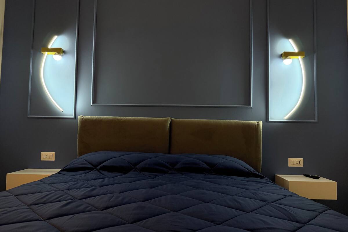 Un lit recouvert d’une couverture matelassée de couleur sombre, avec une tête de lit et deux tables de chevet dotées de lampes.