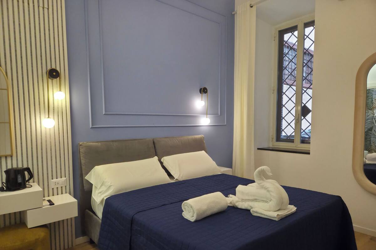 Un lit bien fait, recouvert d’une couverture bleue, une table de chevet avec une bouilloire, et une fenêtre donnant sur un beau paysage.