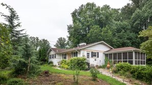 A two-story house with a front porch, a covered porch area, and a garden with various plants and a small tree. - Ground floor apartment 10 mi from D.C. and still feels like countryside! (Bethesda)