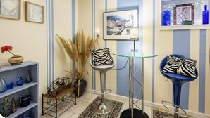 Une pièce aux murs rayés, un tableau encadré, un vase avec une fleur rouge, une étagère en métal avec des vases bleus, une chaise haute avec un coussin à motifs zébrés et une fenêtre avec des rideaux bleus.