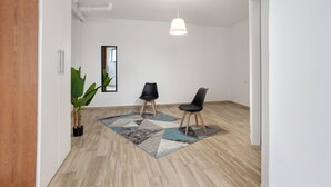 Et værelse med trægulv, to sorte stole, en stor stueplante, et geometrisk tæppe og en lampe samt et spejl på væggen.