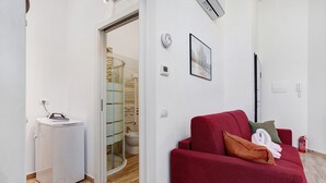 Un espacio habitable compacto con un sofá rojo, un electrodoméstico blanco pequeño, una puerta de cristal que da al baño y un cuadro en la pared.