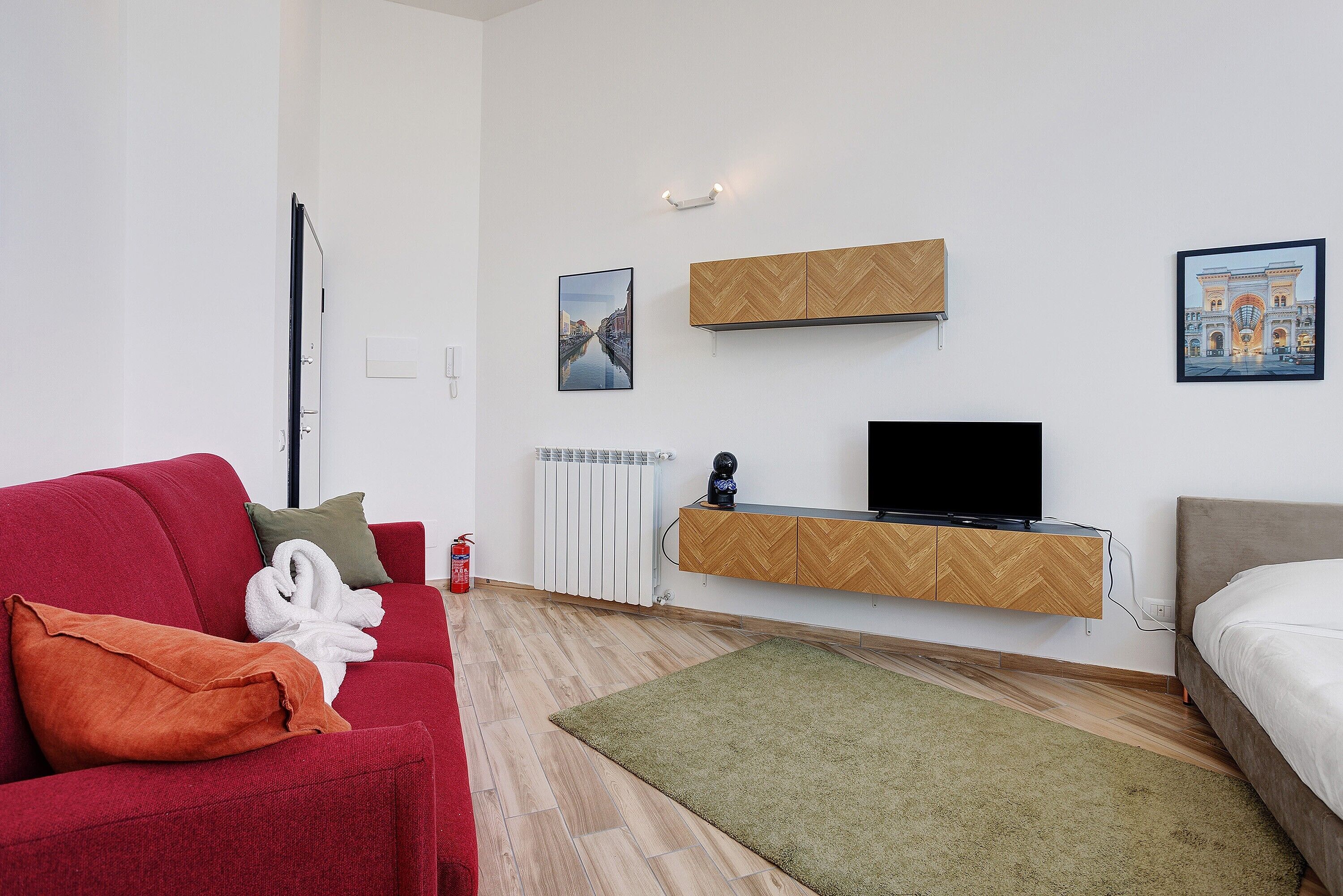 A modern room with a red sofa, a green rug, a bed, a TV on a wooden shelf, and two framed pictures on the wall.