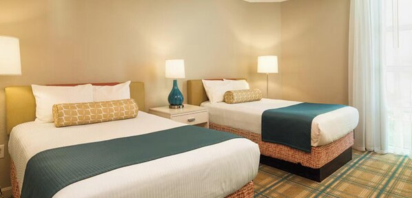 Une chambre d’hôtel avec deux lits, chacun recouvert d’une courtepointe blanche et d’une couverture turquoise. Il y a deux tables de chevet avec des lampes, et la chambre est dotée d’un tapis à carreaux.