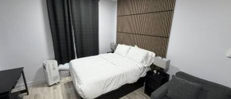 Un dormitorio con cama, un sofá gris, una mesita de noche, un radiador en la pared y una ventana con cortinas.