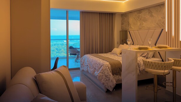 A modern hotel room with a bed, a desk with a chair, a sofa, and a balcony with a view of the city. - Apartamento de lujo Cartagena Beach, Cartagena de indias -Colombia (Cartagena de Indias)