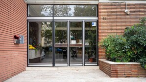 L’entrée d’un immeuble se caractérise par de grandes portes vitrées, un mur en briques et un petit espace jardin agrémenté de plantes en pot.