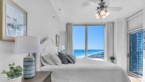 A bedroom with a large bed, a ceiling fan, a view of the ocean, a framed picture on the wall, and a nightstand with a lamp and plant. - Portofino Island Resort 1-1502 (Pensacola Beach)
